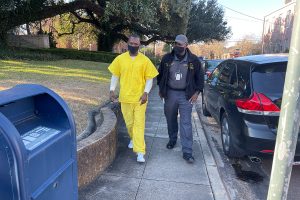 Man in yellow jumpsuit being escorted down a sidewalk by a guard