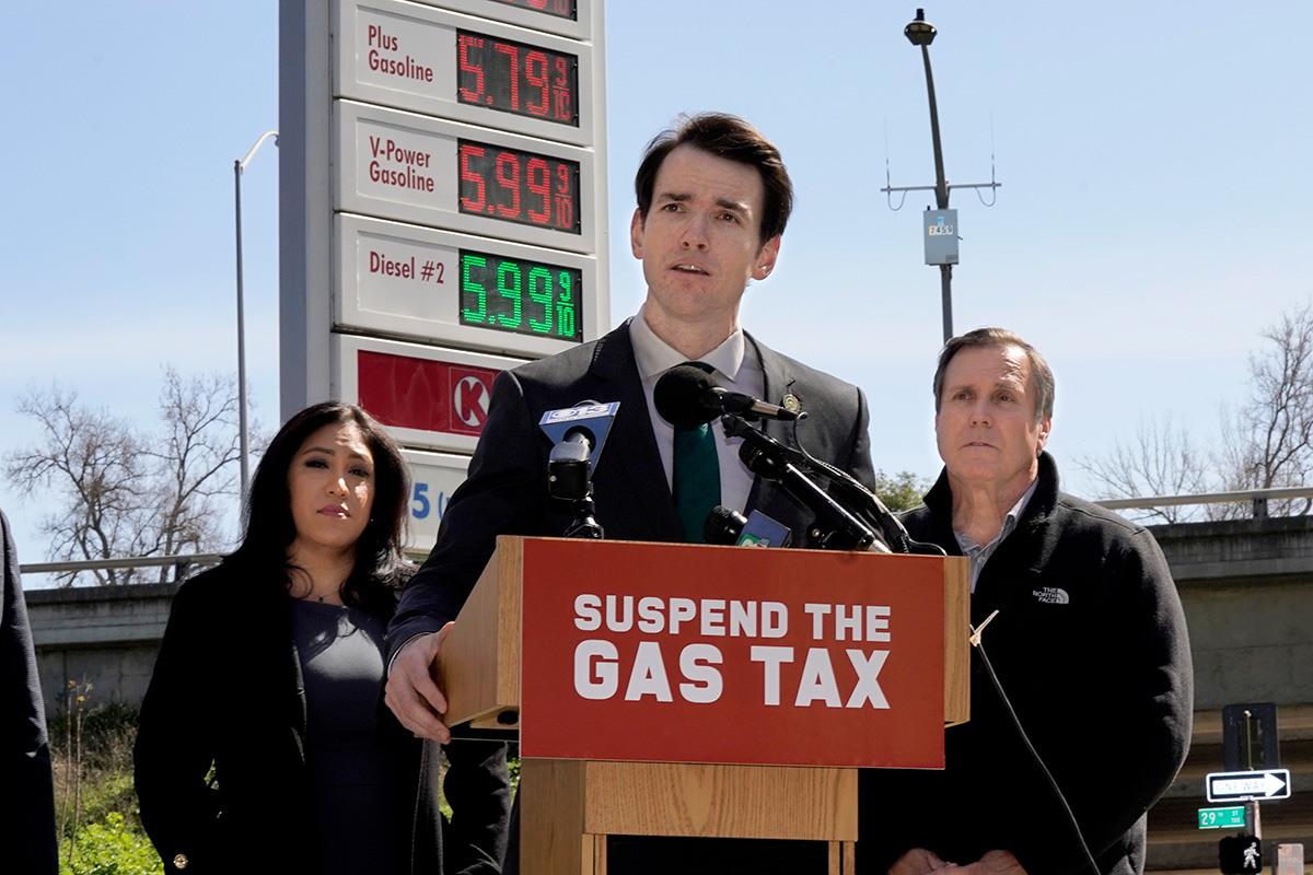 A man speaks at a podium labeled "Suspend the Gas Tax". Two people stand on either side of him. A gas sign with high prices is behind the people.