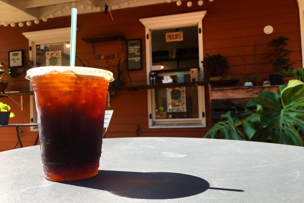 A shot of an iced coffee in a clear to-go cup casting a shadow on a table. The coffee shop is blurred in the background.