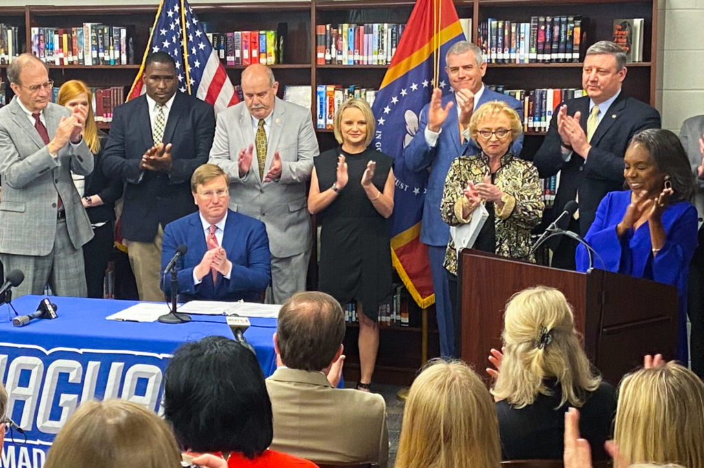 a photo of Governor Tate Reeves signing the teacher pay bill into law while lawmakers and educators standing around him clap