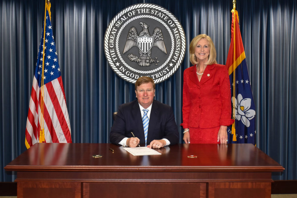 Tate Reeves sits at a desk in front of the Mississippi state seal with Attorney General Lynn Fitch next to him, the two standing between an American flag and Mississippi state flag