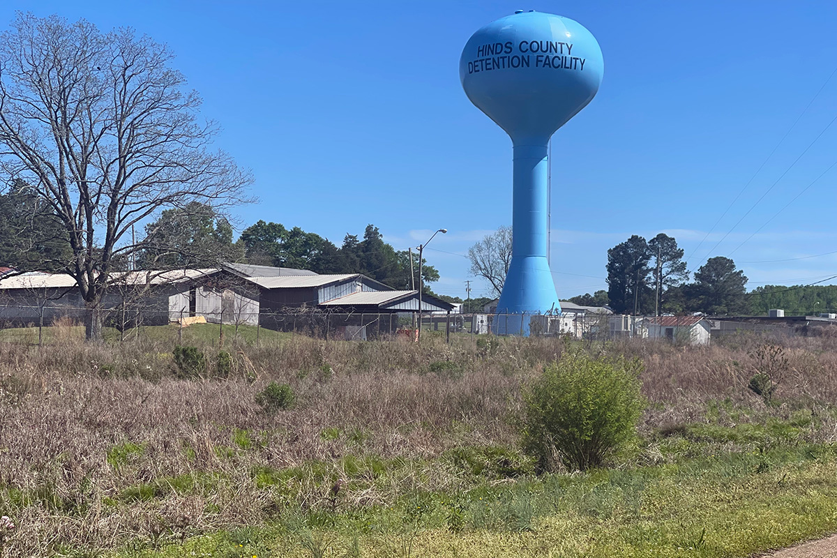 A field with buildings in the background, and a blue water tank that says Hinds County Detention Facility