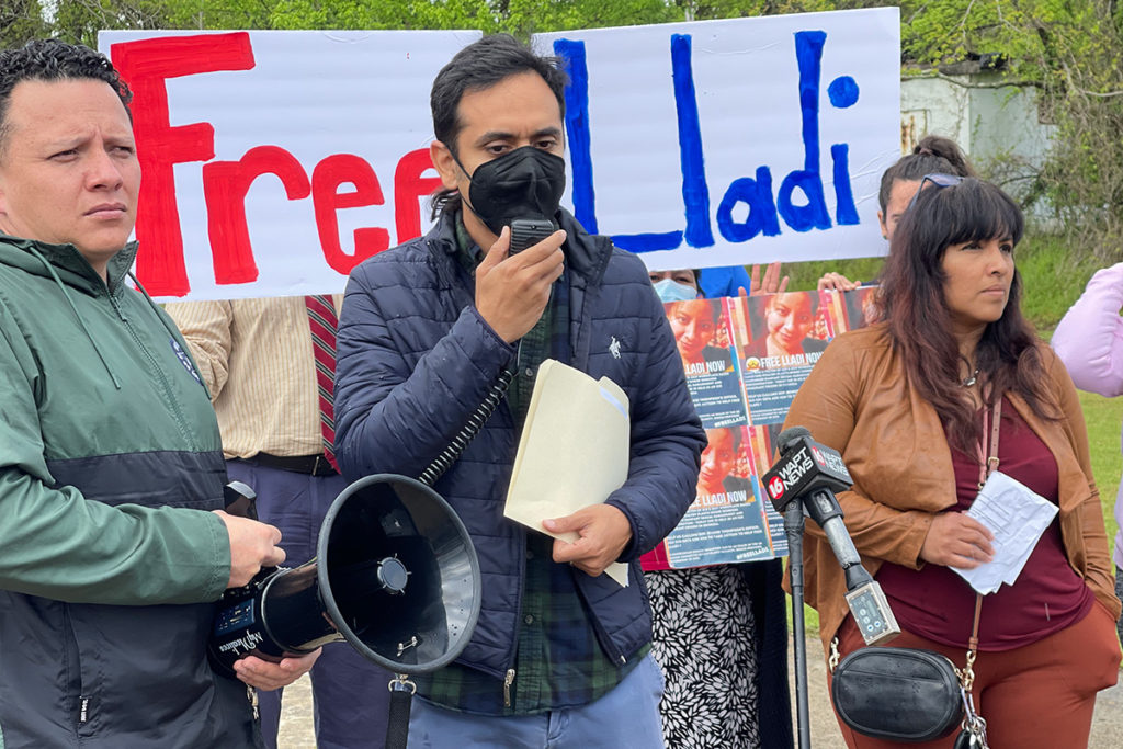 Three people speak with a megaphone in front of a sign that says Free Lladi