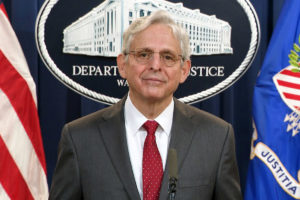 A man in a grey suit and red tie stands between flags and in front of a sign that says Department of Justice