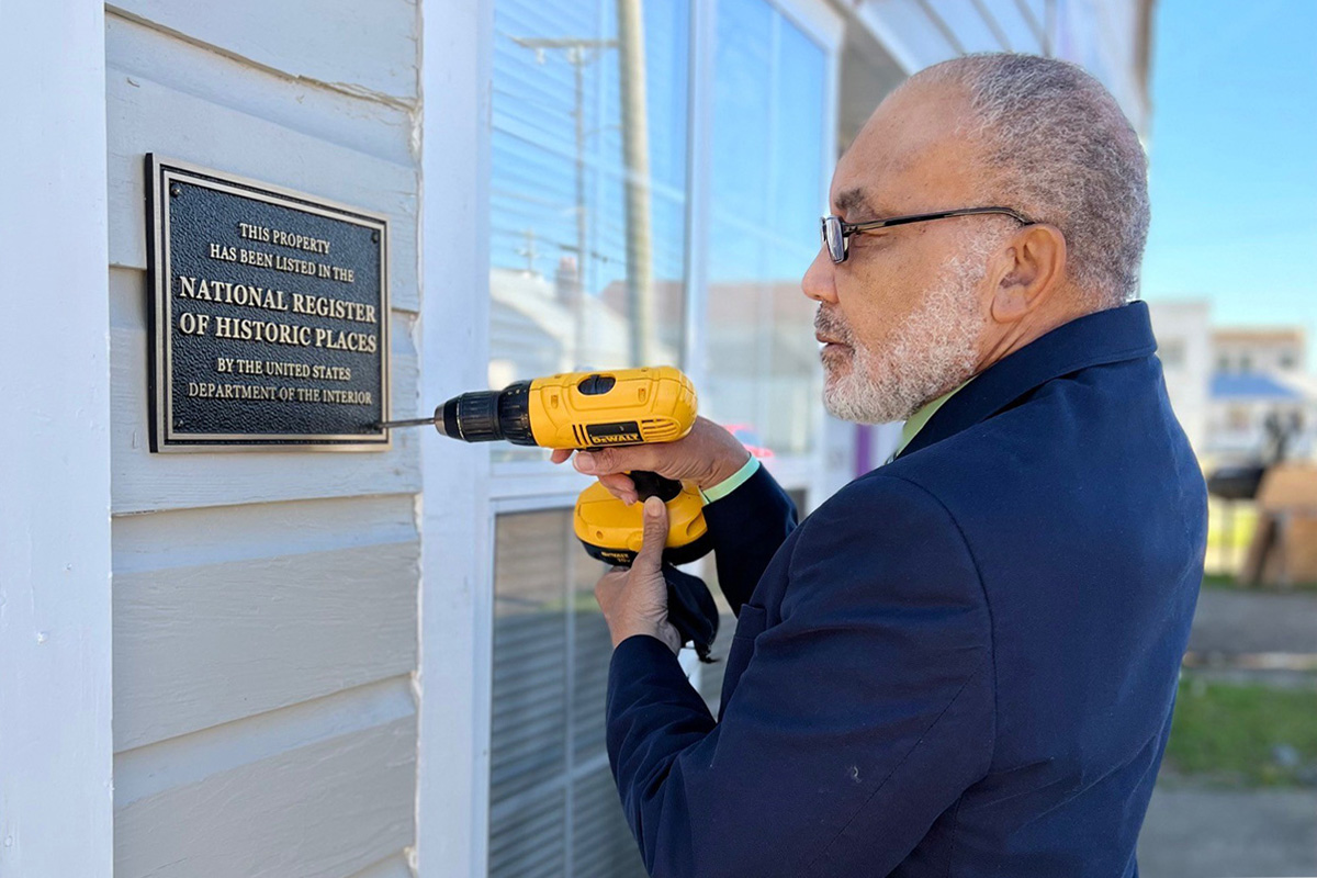 A man in a navy suit is holding a yellow drill to attach a sign to the outside of a beige building. The sign says National Register of Historic Places