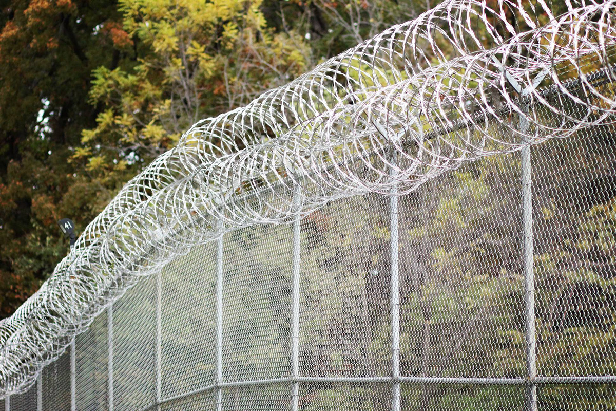 A shot of a chain. link fence with barb wire on top, with trees behind