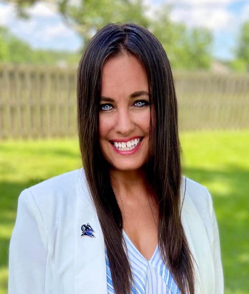 a photo of a woman with long brown hair and blue eyes wearing a white coat with a Blue Devils pin