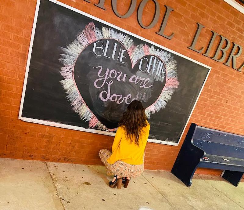 a photo shows a woman stooped down writing the words, 'You Are Loved' on a chalkboard with a colorful heart surrounding the words
