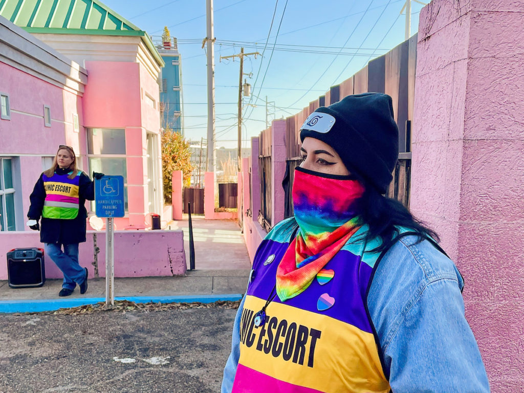 a photo shows a woman in a pink, blue and green mask wearing a vest that says "CLINIC ESCORT" while standing outside the pink painted gate of a pink building with a green roof.