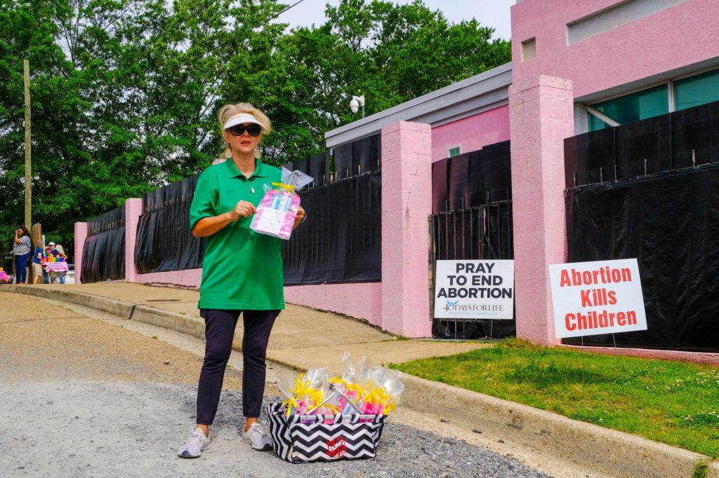 A woman wearing a visor and sunglasses holds a pink and blue bag tied with a yellow ribbon over a basket of other bags in front of a pink building. Signs in the ground read, "Abortion Kills Children" and "Pray to End Abortion"