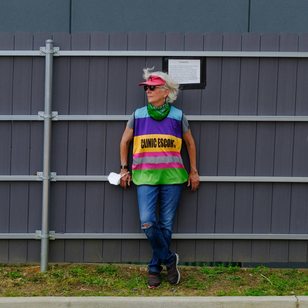 a woman wearing a colorful vest that says "Clinic Escort" stands against a fence, her gray hair blowing in the breeze outside the abortion clinic