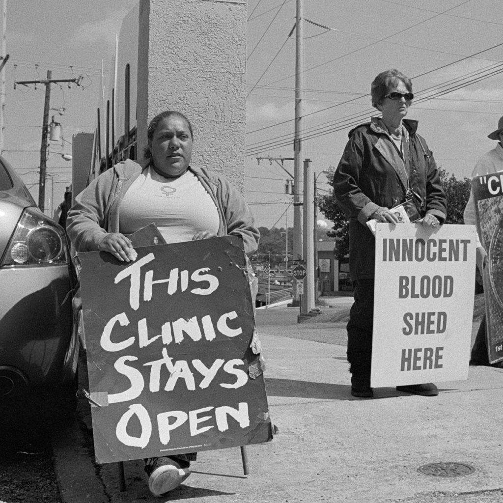 a woman sits holding a sign that says 'This clinic stays open' next to a woman holding a sign that says 'innocent blood shed here' outside the abortion clinic