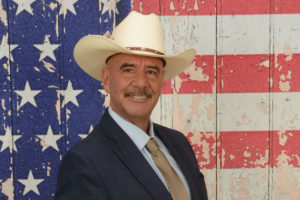 a photo of Mark Strauss, a man in a cowboy hat, smiling in front of an American flag background