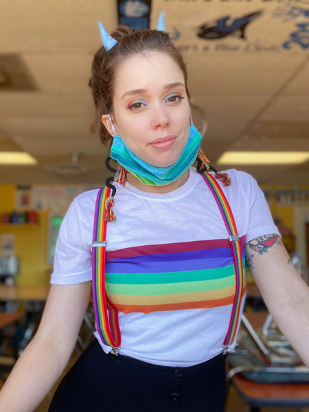 a photo shows Catherine Bass wearing a pair of rainbow suspenders over a white t-shirt with rainbow bars across the center; she is wearing a mask under her chin and blue horns on her head
