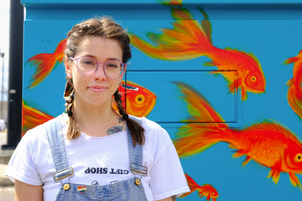a woman with glasses, braided brown pigtails and wearing a pair of overalls atop a white t-shirt poses in front of a painted power box featuring goldfish swimming against a blue backdrop. On her overalls is a small rainbow pin that says, 'Love is Love.'