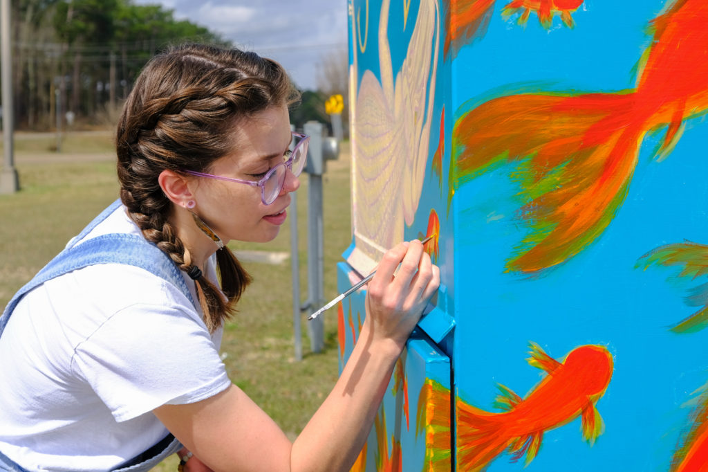 a photo shows Catherine Bass painting goldfish on the side of a utility box
