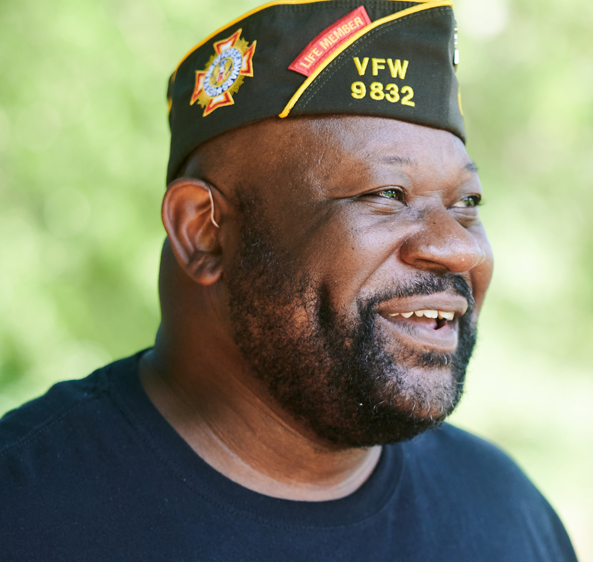 A man wearing a VFW 9832 vet hat smiles to the right