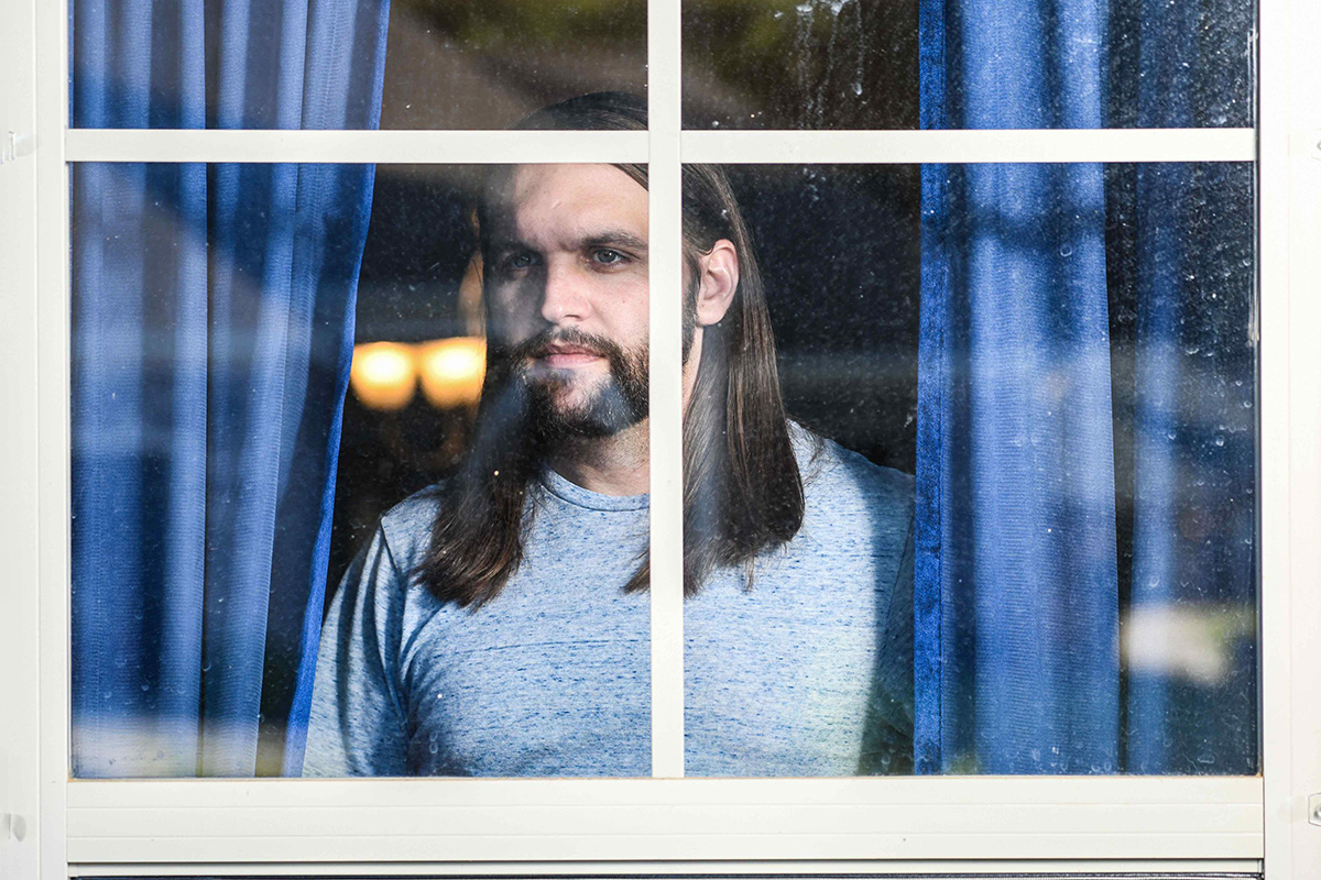 A long-haired man looks out of a window framed by blue curtains