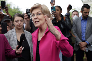 Elizabeth Warren, in a pink jacket, speaks outside in front of a crowd of journalists