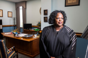 A woman in judge robes stands in the light of a window, her office desk seen behind her