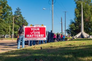 a photo of people standing in a line outside to vote behind a sign that reads, "Your Vote Matters!"