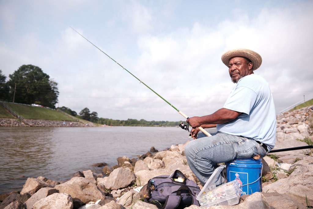 A man sits on a blue bucket on the rocks, fishing
