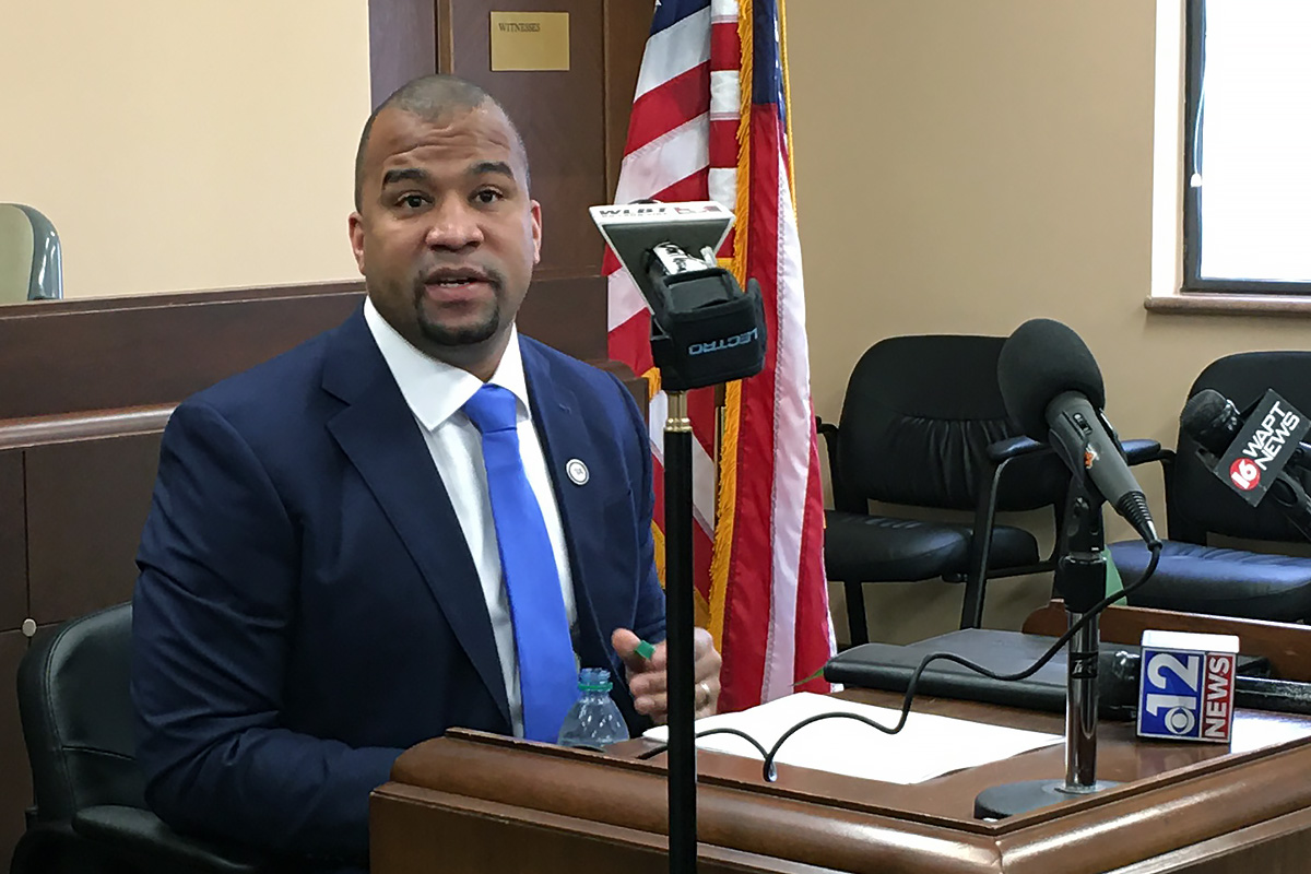 A man in a navy suit and blue tie sits at a desk covered in microphones