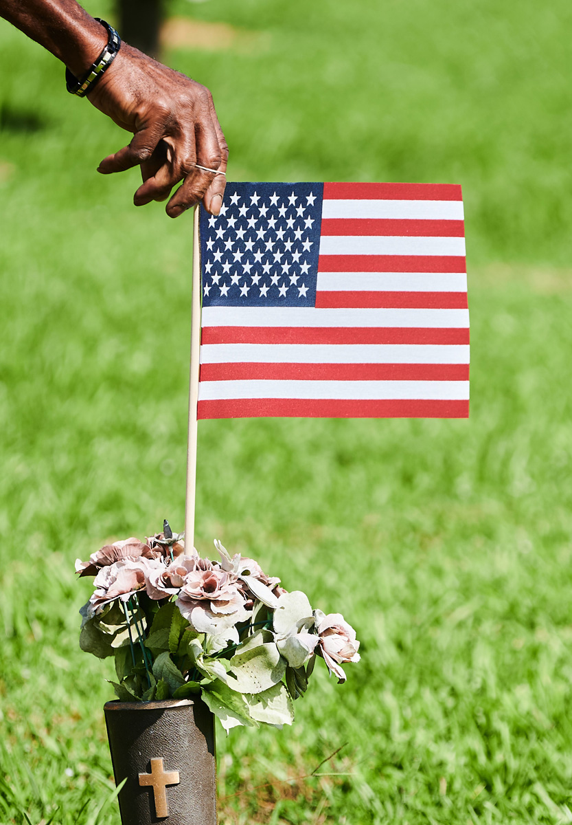 A hand straightens a US Flag in a pot of flowers on a grave