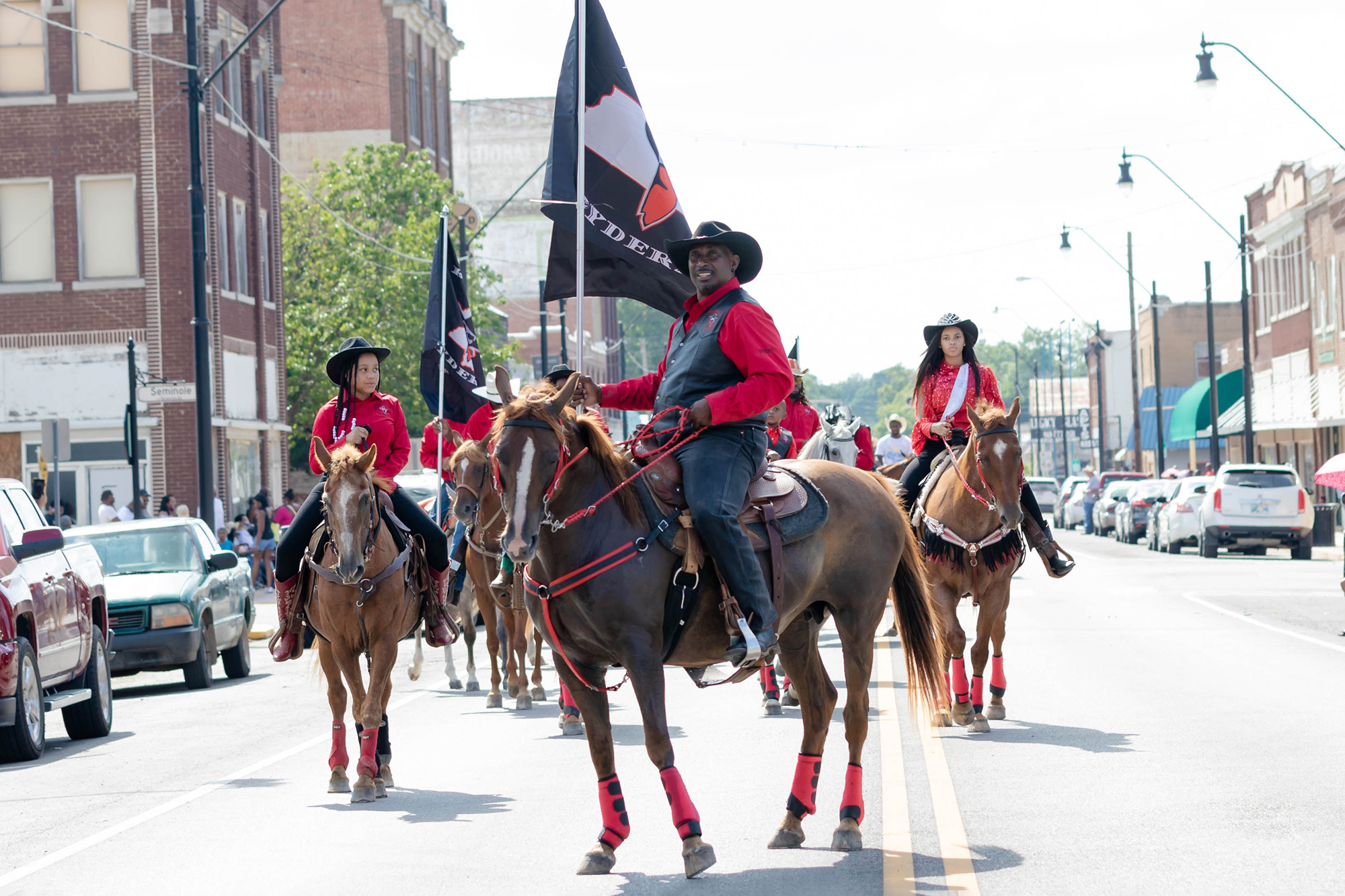 A group of black cowboys wearing black and red, riding brown horses
