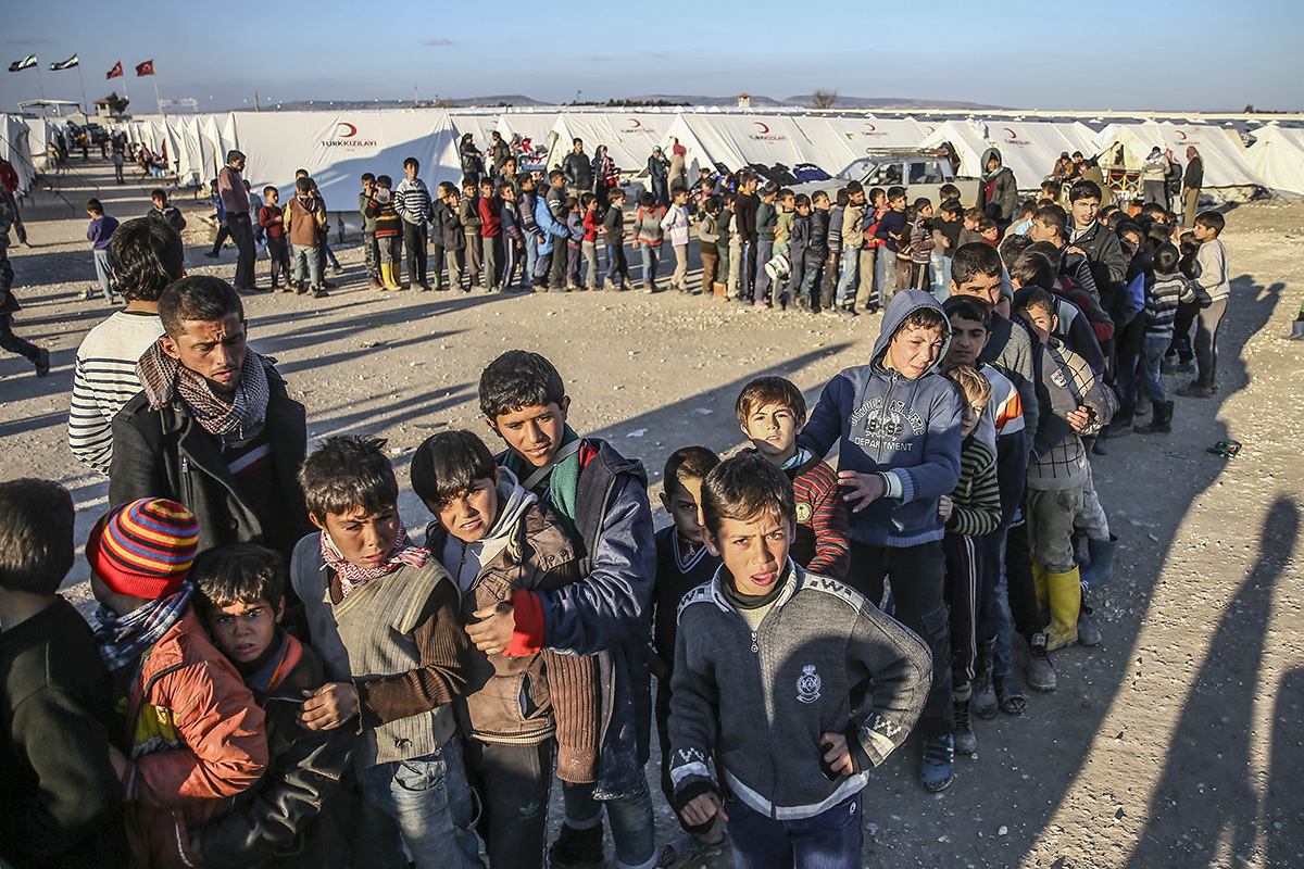 Syrian men and boys line up closely in a long row outside white tents.