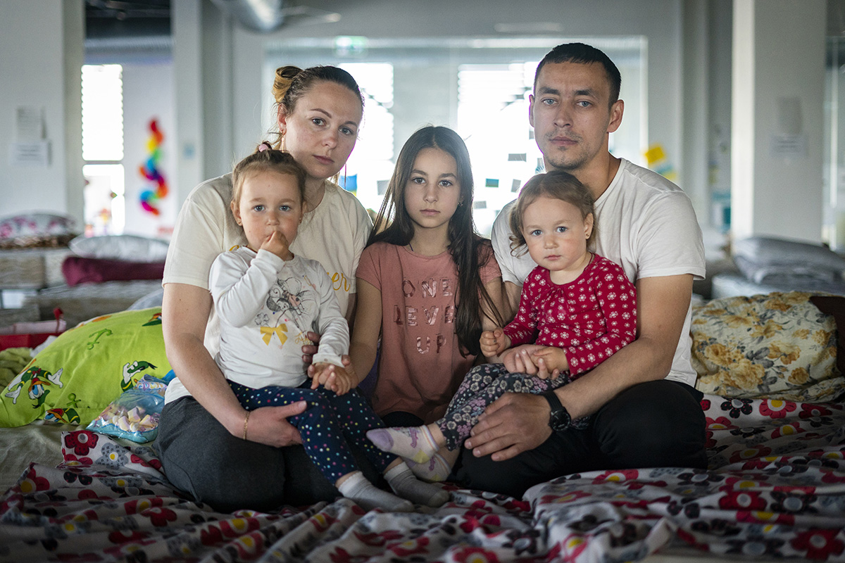 A young woman and man sit on a bed and hold three young children in their laps, looking directly at the camera.