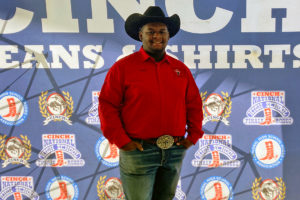 A man in red shirt and black cowboy hat poses in front of a blue sign
