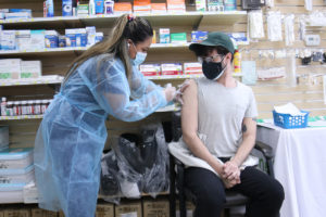 A man getting a vaccine in his arm by a woman in blue PPE. A wall of medical supplies is behind them.