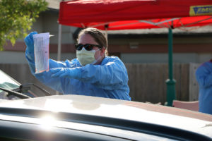 A woman in blue PPE and yellow face mask picks up a plastic bag from a car's windshield in a drive through COVID testing line