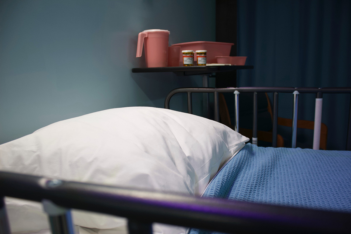An empty hospital bed with blue blanket tucked neatly under a white pillow. A shelf with coral water pitcher and medicine bottles sits above it