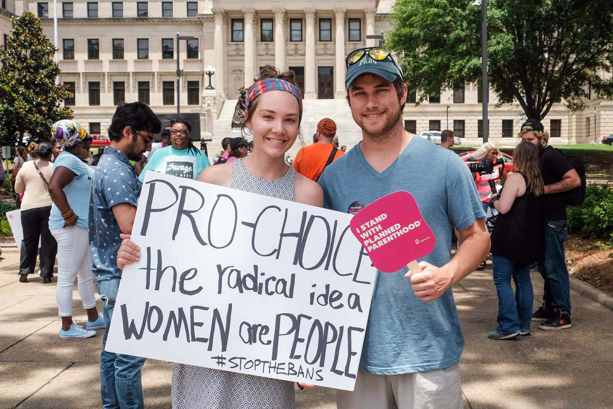Two people hold signs that say "Pro-Choice the radical idea Women are people #stopthebans" and "I stand with Planned Parenthood"