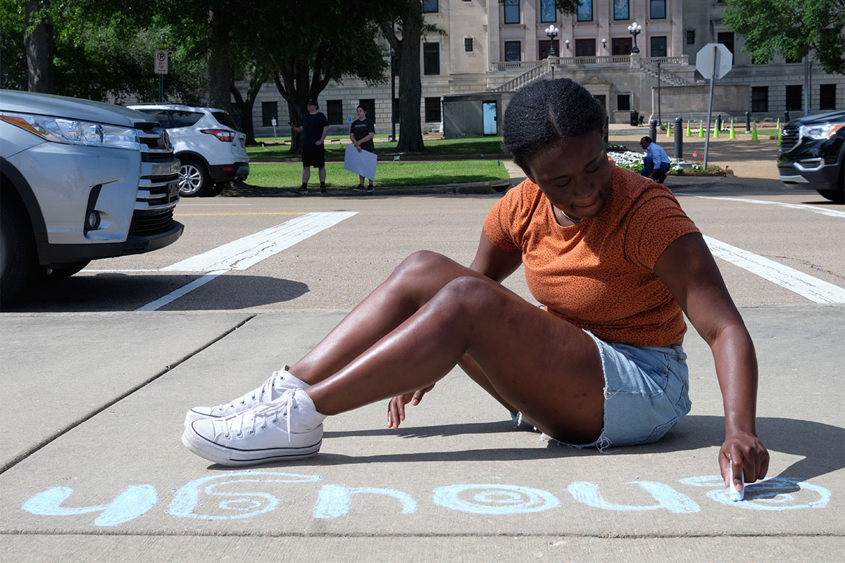 A woman sits on the ground and writes "enough" in charlk on the sidewalk