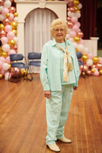 A woman in a mint suit and cream tie stands in the center of a room, balloons on the wall behind her