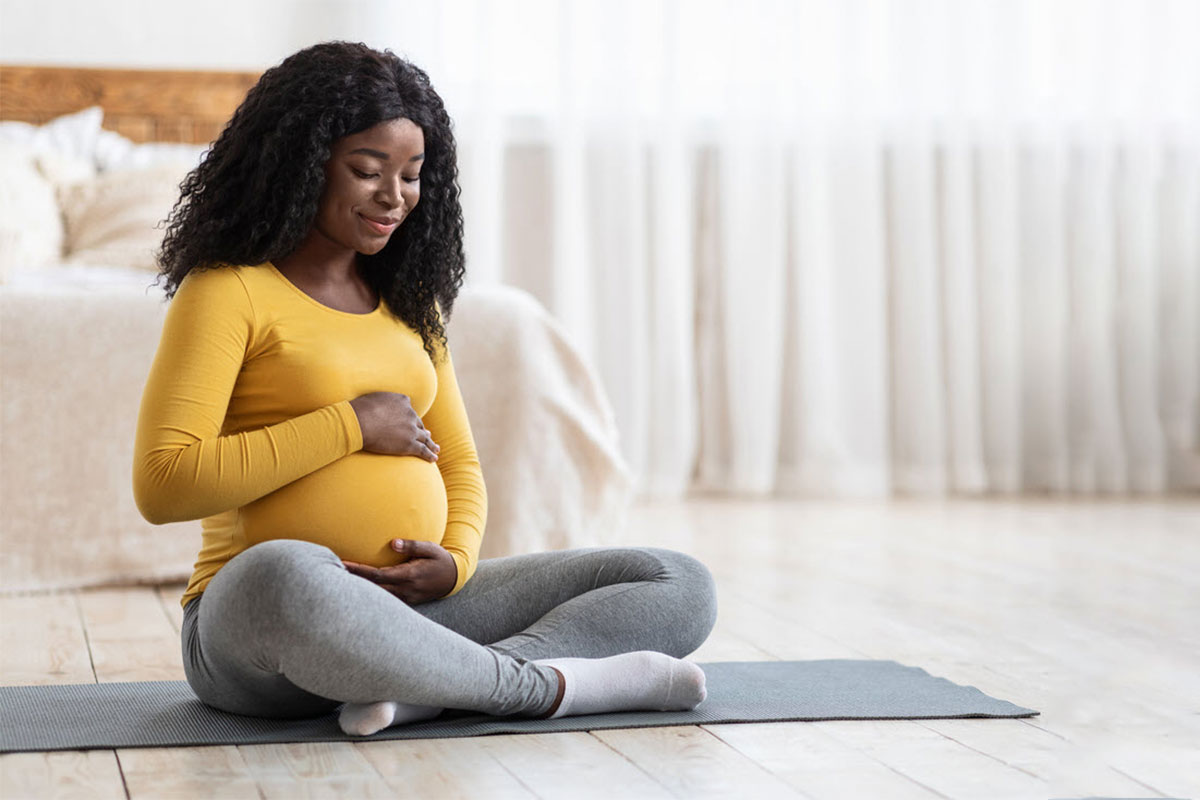 A pregnant woman sits on a grey yoga mat and hugs her belly happily