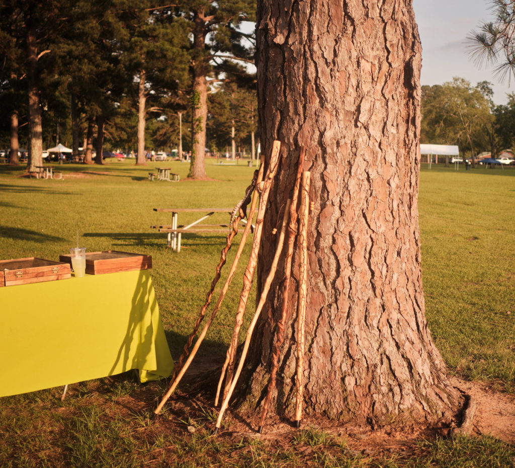 Wooden canes lean against a tall tree trunk, a yellow table beside holds more wooden items