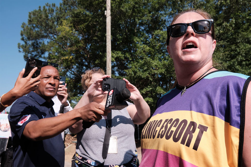 a photo shows a woman wearing a vest that says "clinic escort" while people with cameras stand behind her