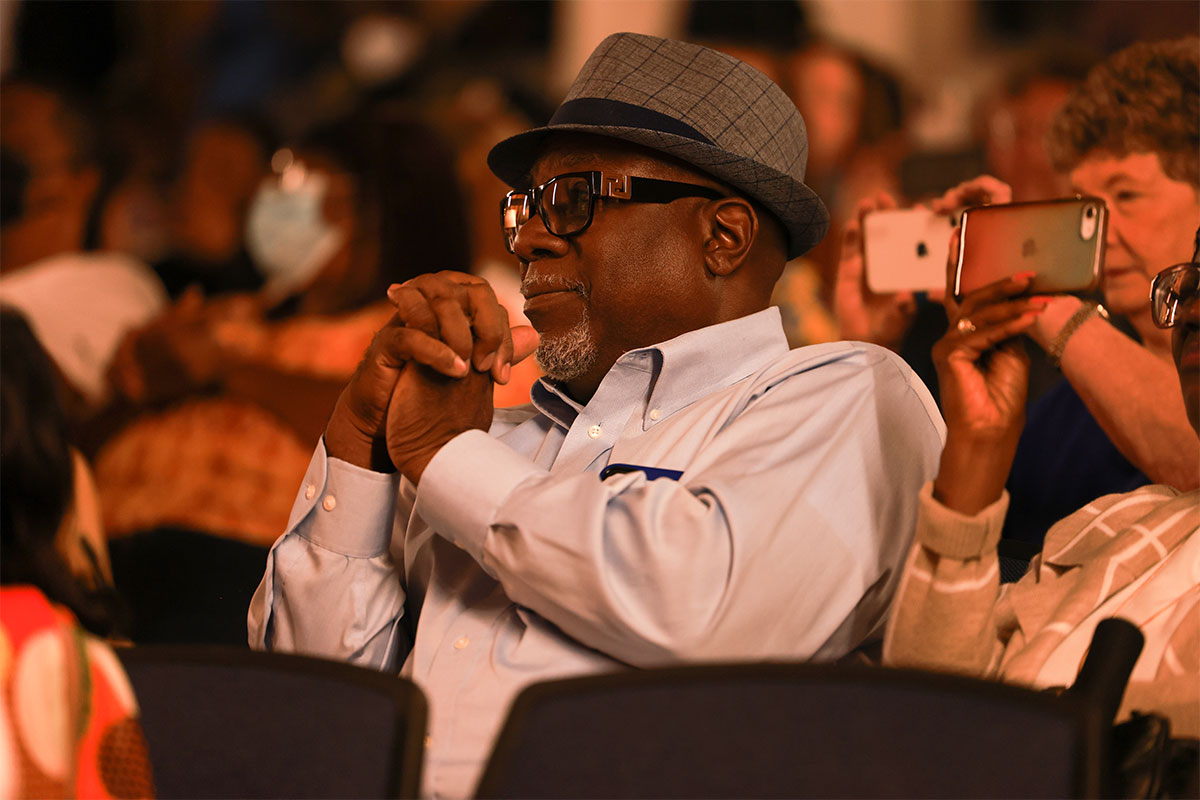 A close up of a man wearing glasses and a hat in a seated crowd