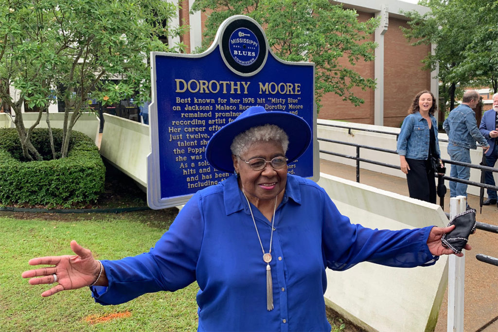 Dorothy Moore, dressed in all blue, stands with arms wide in front of a blue marker with her name