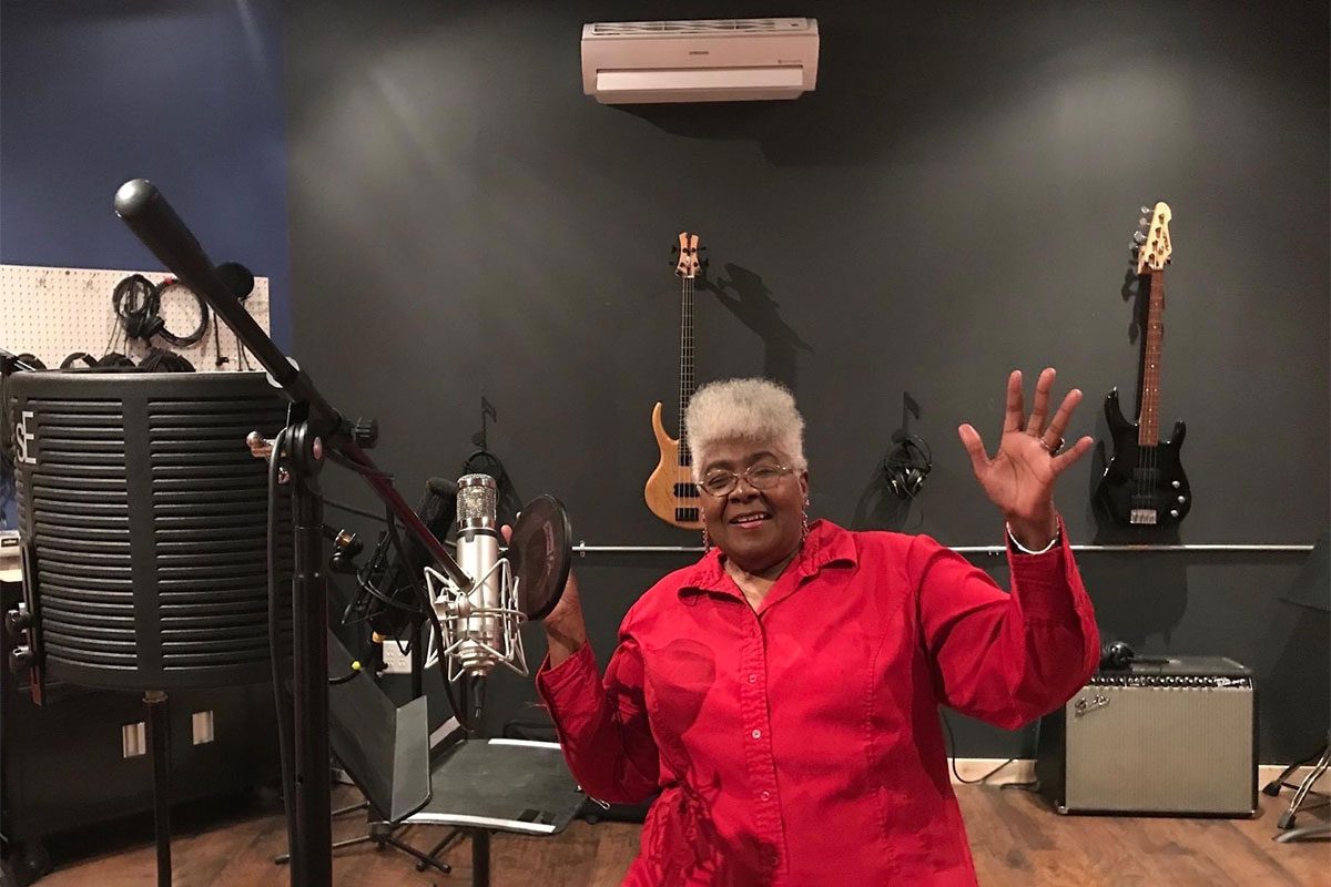 A woman in red sits with hands spread wide in a music studio