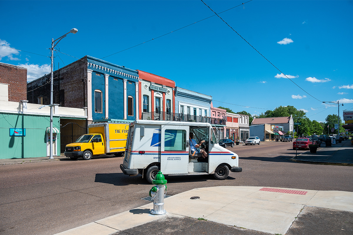 A USPS mail truck is seen on the corner of a old strip of colorful buildings