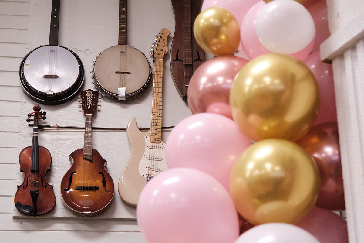 Dulcimers hang on a white wall, pink and gold balloons blurred in the foreground