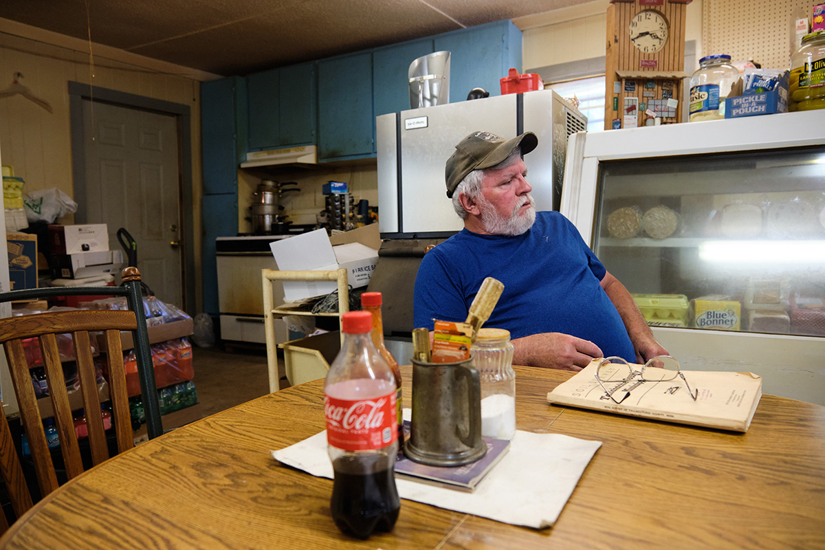 A man in a blue tshirt and green baseball hat leans back at a wooden table in a general store