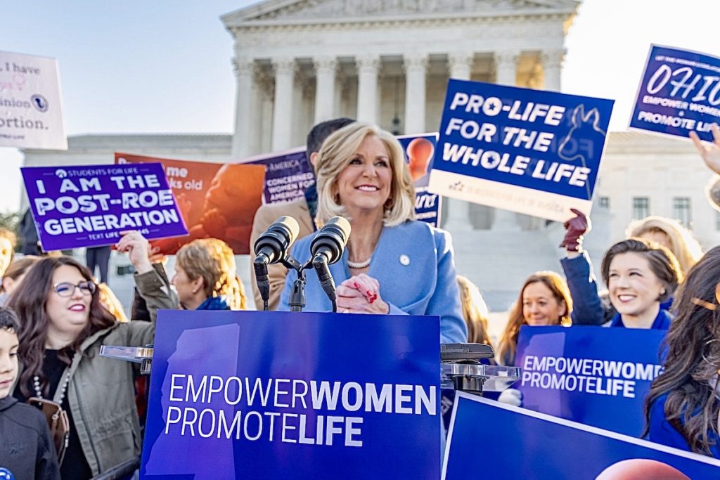 a photo of Lynn Fitch smiling in front of the Supreme Court with people around her holding signs that say things like, "Empower Women, Promote Life," "I Am The Post Roe Generation," and "Pro Life For The Whole Life."