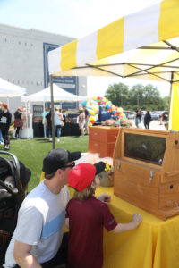 A man and child look at a box on a yellow table under a yellow and white striped tent