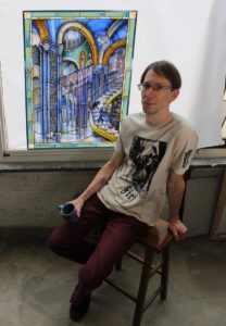 Artist Rob Cooper sits on a stool in front of his stained glass window art